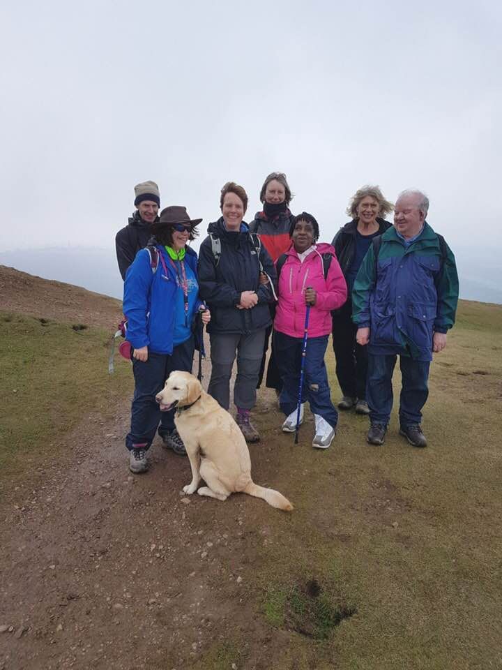 A group of 7 walkers with a guide dog posing for a picture on a hillside path